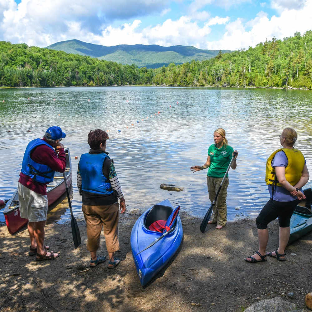four people standing at the edge of a lake with kayaks and paddles